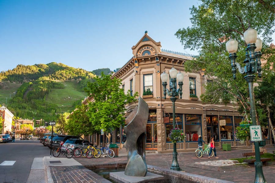 Downtown Aspen in summer: a Victorian brick corner building, street lamps and bikes, with Aspen Mountain behind.