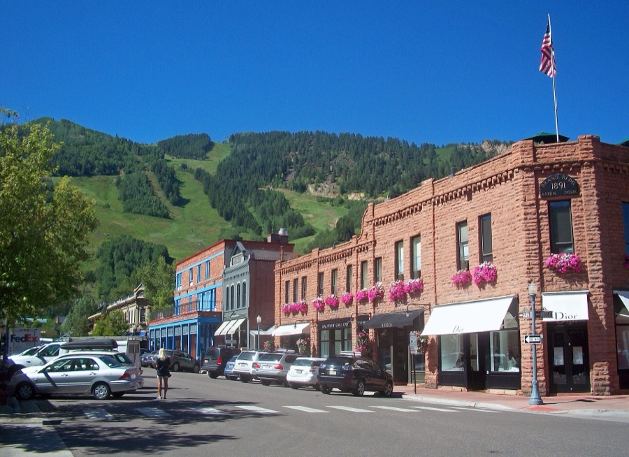 Downtown Aspen in summer, with the green ski runs of Aspen Mountain rising behind the brick storefronts.