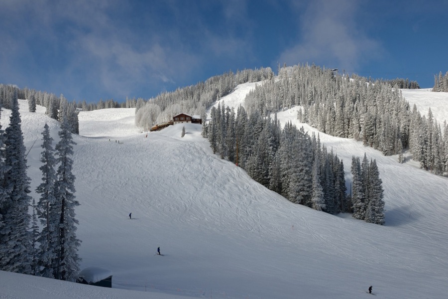 Skiers on a sunlit run at Aspen Mountain after fresh snow, with a lodge among the firs above.