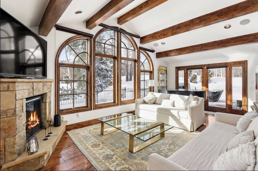 Living room with stone fireplace, timber beams, arched windows looking onto snow, two cream sofas and a grand piano.