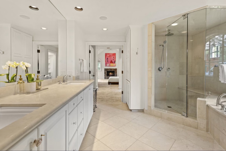 Primary bathroom with a long stone vanity and dual sinks, glass steam shower, jetted tub, and a view through to the bedroom fireplace.
