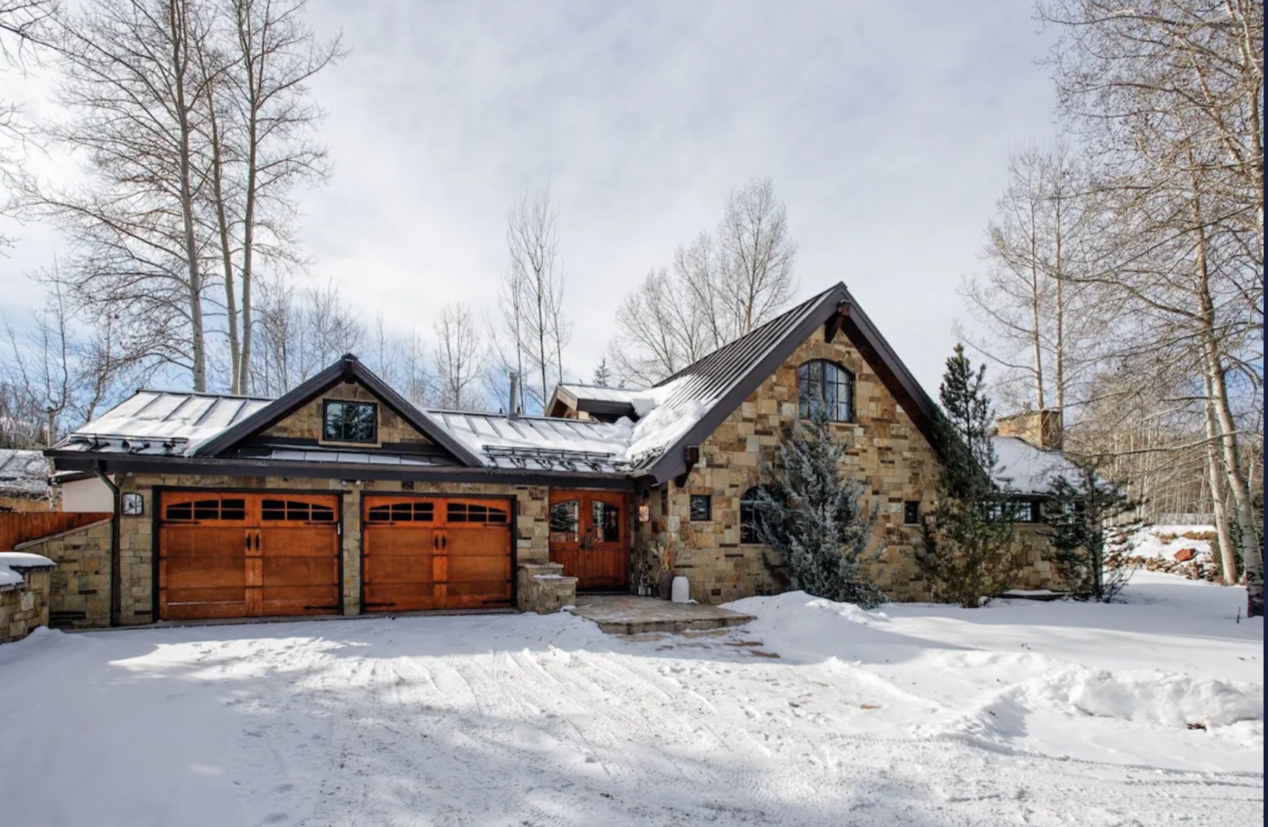 Front of the stone house in winter with two timber garage doors, a snow-covered driveway and aspen trees.