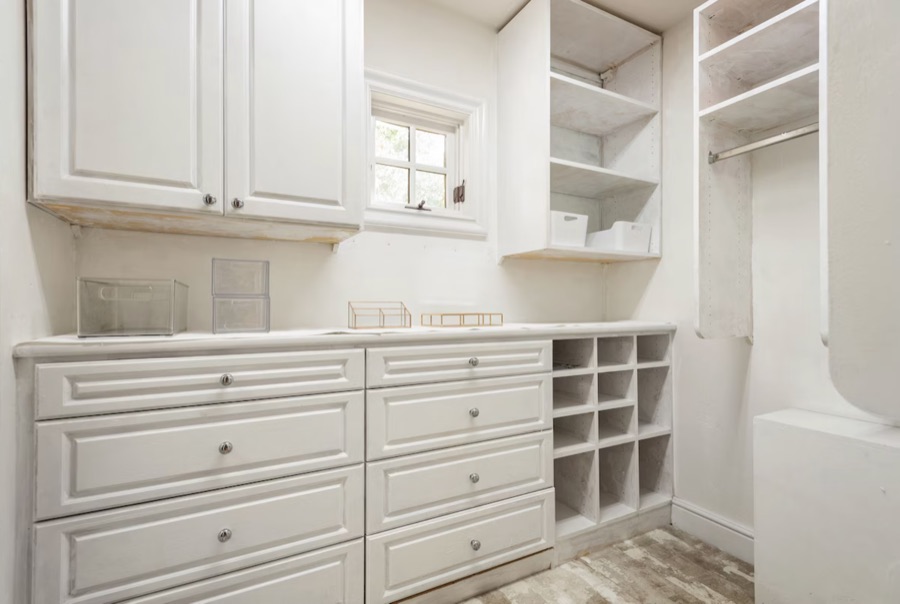Walk-in closet with white built-in drawers, cubbies, hanging rails and a small window.